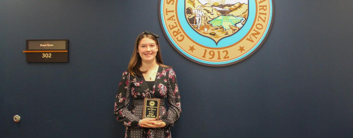 Eliza McCall, one of the February 2025 students of the month holding her award and standing in front of the Arizona State seal
